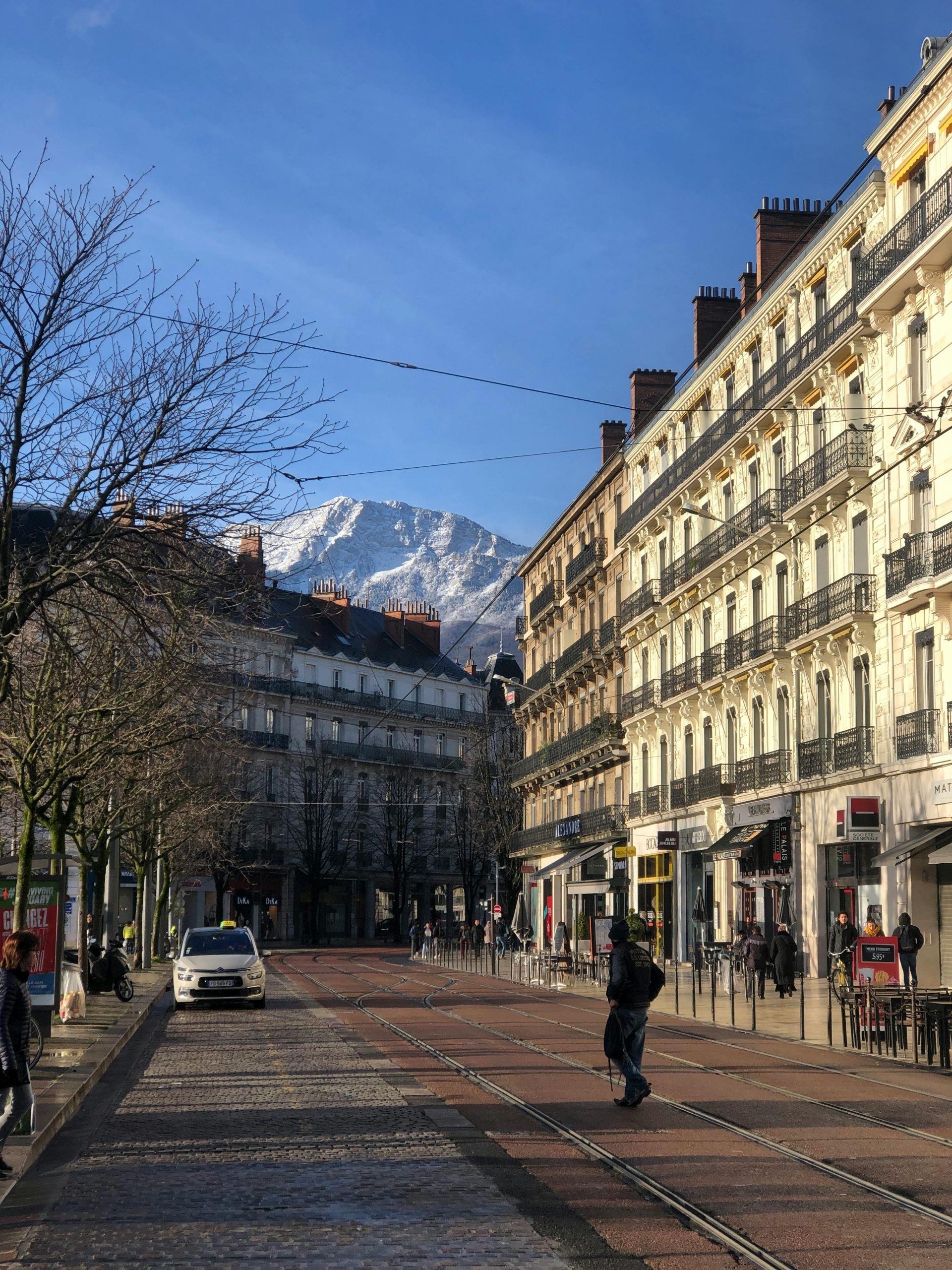 A picturesque street in Grenoble, showcasing historic architecture and a stunning mountain backdrop.